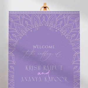 May include: A lavender wedding welcome sign on a wooden easel. The sign features ornate floral designs and the names "Krish Rajput and Ananya Kapoor." Text reads "Welcome to the wedding of" and "We're so glad you're here."