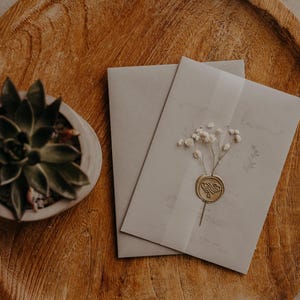 May include: Two white envelopes with a wax seal and a sprig of dried white flowers on a wooden surface. A small potted succulent plant is in the background.