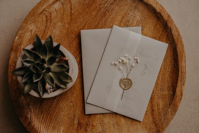 May include: Two white envelopes with a wax seal and a sprig of dried white flowers on a wooden surface. A small potted succulent plant is in the background.