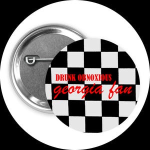 Puede incluir: Un botón redondo con un patrón de cuadros blancos y negros. El botón tiene las palabras "DRUNK OBNOXIOUS georgia fan" en texto rojo. La parte posterior del botón es plateada.