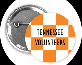 Könnte beinhalten: Ein runder Button mit silbernem Metallrücken und einem orange-weiß karierten Muster. Der Text "TENNESSEE VOLUNTEERS" ist in Schwarz dargestellt. Der Button ist vor einem weißen Hintergrund platziert.
