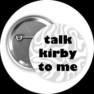 Puede incluir: Un botón redondo con una parte trasera de metal plateado y un diseño en espiral blanco y gris. El botón presenta el texto "talk kirby to me" en letras negras en negrita.