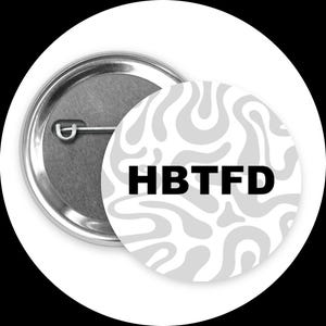 Puede incluir: Un botón redondo con una parte trasera de metal plateado y una cara blanca. La cara tiene un patrón gris con remolinos y las letras negras "HBTFD". El botón está sobre un fondo blanco.