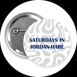 May include: A round button with a silver metal back and a white face. The face has a gray, swirled pattern and the text "SATURDAYS IN JORDAN-HARE" in navy blue.
