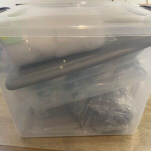 May include: A clear plastic storage bin with a blue lid, filled with various items. The contents are partially visible, including what appears to be a dark-colored object and other wrapped items. The bin is on a wooden surface.