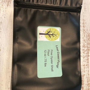 May include: A black resealable bag with a white label that says "Lost Coast Fungi Fine Oyster Shell Flour 12 oz / .75 lbs" and a green illustration of a tree with white hearts.