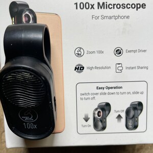 May include: A black 100x microscope for smartphones. The device has a clip to attach to a phone and features high-resolution capabilities. The packaging includes instructions for easy operation.