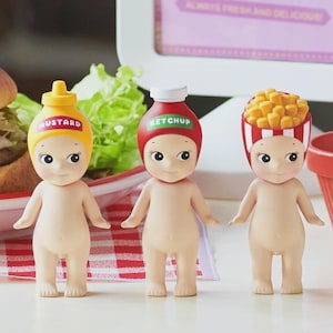 May include: Three small, nude-colored figurines with food-themed hats: mustard, ketchup, and french fries. The mustard hat is yellow, the ketchup hat is red, and the fries hat is red and white striped. A sign in the background reads "Burger in Town."