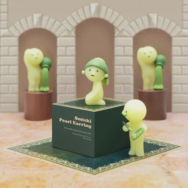 May include: A display of Smiski figurines. The central figure, wearing a green cap, sits on a dark green box labelled "Smiski Pearl Earring." Other figures stand on pedestals, and one holds a small object. The background features arched structures.