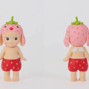 May include: A small figurine of a character wearing a pink strawberry-patterned hat with a green stem on top and red strawberry-patterned shorts. The figure has a light skin tone, dark eyes, and small white wings on its back, shown from front and back.