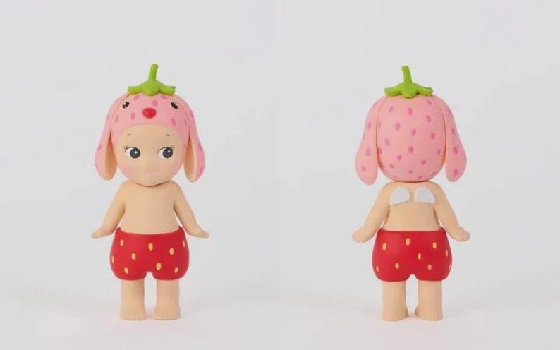 May include: A small figurine of a character wearing a pink strawberry-patterned hat with a green stem on top and red strawberry-patterned shorts. The figure has a light skin tone, dark eyes, and small white wings on its back, shown from front and back.