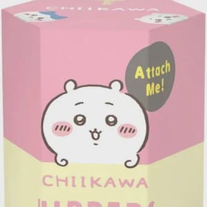 May include: A white Chiikawa figure sits atop a pink and yellow box. The box features a cartoon illustration of the character with the text "CHIIKAWA HIPPERS DECORATIVE MINI FIGURES". A gray speech bubble says "Attach Me!"