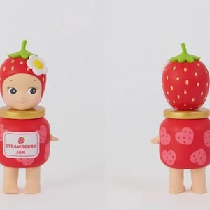 May include: A small figurine featuring a character wearing a red strawberry hat with a white flower, standing atop a red jar-shaped body. The front label reads STRAWBERRY JAM with a small strawberry icon, while the back shows pink heart-shaped strawberries.