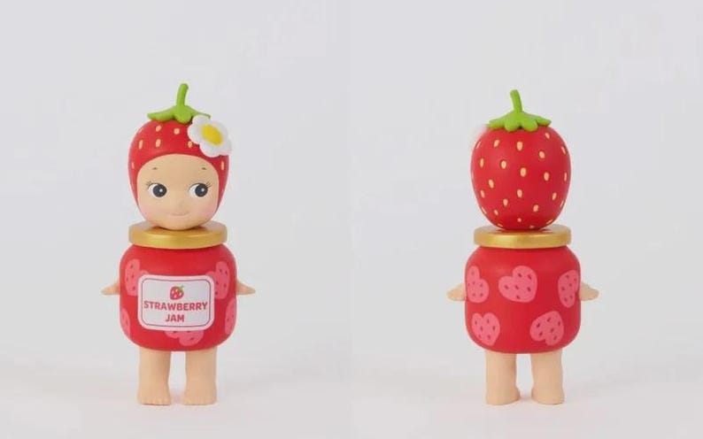 May include: A small figurine featuring a character wearing a red strawberry hat with a white flower, standing atop a red jar-shaped body. The front label reads STRAWBERRY JAM with a small strawberry icon, while the back shows pink heart-shaped strawberries.