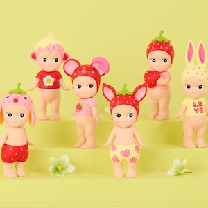 May include: A collection of six small collectible figurines with pale skin, each wearing a unique headpiece or hat featuring strawberry, floral, or animal-themed designs, and some wearing small shirts or shorts with heart and flower patterns.
