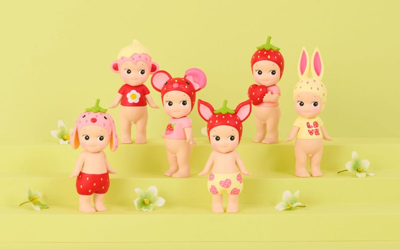 May include: A collection of six small collectible figurines with pale skin, each wearing a unique headpiece or hat featuring strawberry, floral, or animal-themed designs, and some wearing small shirts or shorts with heart and flower patterns.