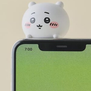 May include: A white, round, cartoon-style figure with a smiling face, pink cheeks, and small ears sits atop a smartphone. The phone screen displays a green background and the time 7:00.