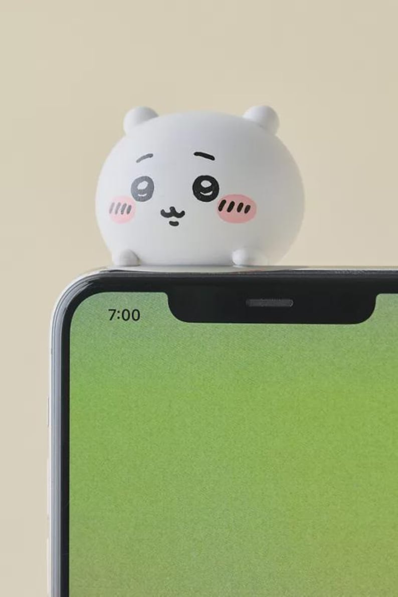 May include: A white, round, cartoon-style figure with a smiling face, pink cheeks, and small ears sits atop a smartphone. The phone screen displays a green background and the time 7:00.
