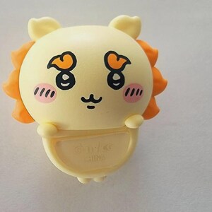 May include: A light yellow cartoon character with orange accents, blush, and a smiling face. The character has a round head with small ears and a small body. The character is holding a small, light yellow, semi-circular object.
