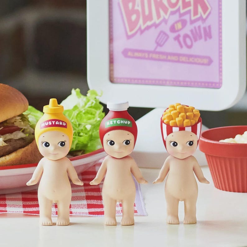 May include: Three small, nude-colored figurines with food-themed hats: mustard, ketchup, and french fries. The mustard hat is yellow, the ketchup hat is red, and the fries hat is red and white striped. A sign in the background reads "Burger in Town."