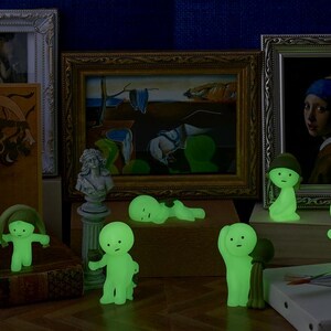 May include: A display of luminous green figurines in different poses, set before framed artworks and decorative items. The figurines have simple, rounded features and glow, creating a whimsical and artistic arrangement.