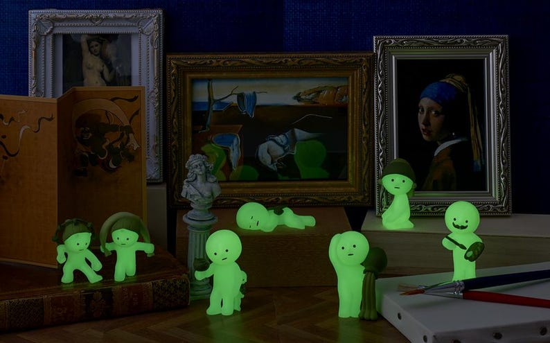 May include: A display of luminous green figurines in different poses, set before framed artworks and decorative items. The figurines have simple, rounded features and glow, creating a whimsical and artistic arrangement.