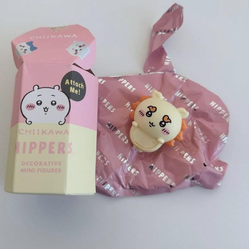 May include: A pink and cream-colored box with the text "CHIIKAWA HIPPERS" and a cartoon character illustration. Next to it is a small, cream-colored figure with orange accents, packaged in pink foil. The foil is printed with the word "HIPPERS".