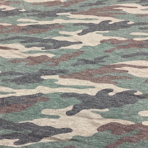 May include: Close-up of a camouflage pattern fabric. The design features irregular shapes in shades of green, brown, gray, and beige. The fabric appears to be a soft, knit material, suitable for clothing or accessories.
