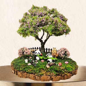 May include: A miniature garden scene with a small tree with pink flowers, two panda bears, a small owl, and a wooden fence. The garden is surrounded by moss and small flowers.