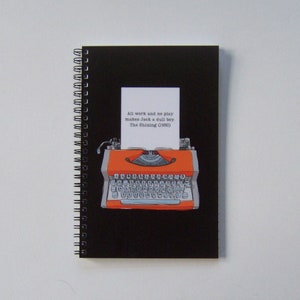 May include: A black spiral bound notebook with a red and black typewriter illustration. The notebook has a white paper page with the text "All work and no play makes Jack a dull boy. The Shining (1980)"