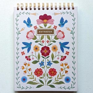 May include: A spiral-bound notebook with a floral design. The cover features a symmetrical pattern of flowers, leaves, and blue birds on a white background. The word "NOTEBOOK" is in a brown rectangle. The spiral binding is gold.