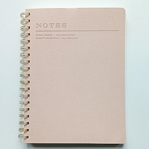 May include: A blush-colored spiral-bound notebook with the word "NOTES" embossed on the cover. The notebook has a gold spiral binding and includes the text "SPIRAL BOUND 1 192 LINED PAGES QUALITY MATERIALS L No. FSS42000".
