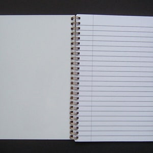Pode incluir: Um caderno de espiral aberto com p&aacute;ginas pautadas &agrave; direita e p&aacute;ginas em branco &agrave; esquerda. O caderno tem uma encaderna&ccedil;&atilde;o prateada.