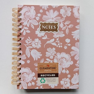 May include: A spiral-bound notebook with a floral pattern in white on a dusty rose background. The cover features the word "NOTES" in a gold-colored label. The notebook is labeled "RECYCLED" and made with 60% recycled fiber.