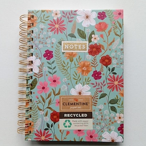 May include: A spiral-bound notebook with a floral design on a teal background. The cover features pink, orange, and white flowers with green leaves. The notebook has gold spiral binding and the word "NOTES" is printed on the cover. The notebook is made with recycled paper.