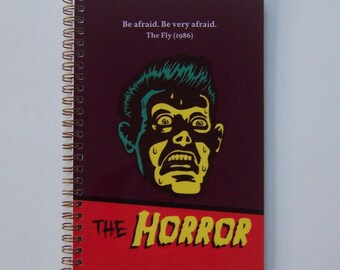Pode incluir: Um caderno roxo com encadernação em espiral. A capa apresenta uma ilustração amarela e verde de um rosto de homem com uma expressão de horror. O texto "Be afraid. Be very afraid. The Fly (1986)" está impresso em branco na parte superior da capa. O texto "THE HORROR" está impresso em amarelo na parte inferior da capa.