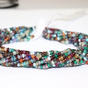 May include: A strand of colourful gemstone beads, including green, blue, red, and brown, arranged in a circle on a white surface.