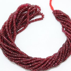 May include: A strand of deep red ruby beads, each bead is faceted and has a slightly irregular shape. The beads are strung on a thin thread and are arranged in a loose, flowing pattern.