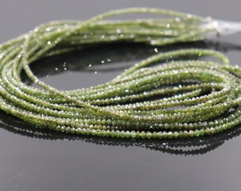 May include: A strand of green diamond beads. The beads are small and round, and they have a faceted finish. The strand is long and is lying on a gray surface.