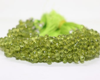 May include: A collection of faceted, olive-green gemstone beads. The beads are strung together, forming a cluster. A bundle of bright green string is visible in the background, tied with a yellow band. The beads are on a white surface.