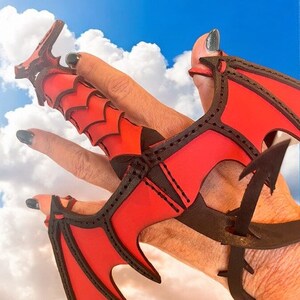 May include: A red leather dragon claw ring with black stitching. The ring is worn on a hand with black nail polish.