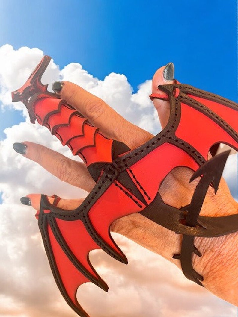 May include: A red leather dragon claw ring with black stitching. The ring is worn on a hand with black nail polish.