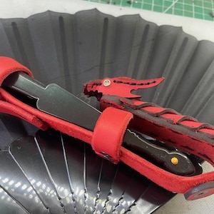 May include: A red leather dragon-shaped sheath holding a black straight razor. The sheath has a dragon head with a rhinestone eye. The razor is black with a gold-colored dot on the handle. The sheath is on a black fan.