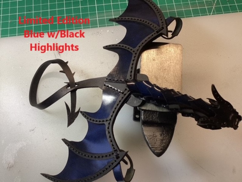 May include: A blue leather dragon wing with black highlights. The wing is attached to a black leather strap. The strap has a loop at the end. The text "Limited Edition Blue w/Black Highlights" is visible in the image.