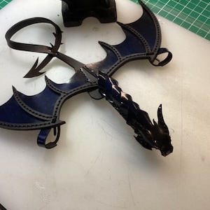 May include: A black leather dragon with blue wings. The dragon is made of leather and has a detailed design. The wings are spread out and the dragon's head is facing forward.