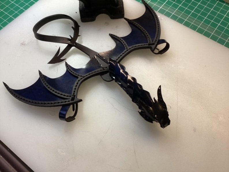 May include: A black leather dragon with blue wings. The dragon is made of leather and has a detailed design. The wings are spread out and the dragon's head is facing forward.