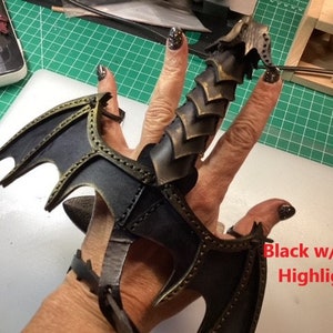 May include: Black leather dragon claw ring with gold accents. The ring is worn on a finger and has a wing design on the side. "Black w/ Gold Highlights" is written on the image.