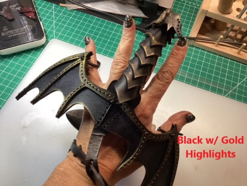 May include: Black leather dragon claw ring with gold accents. The ring is worn on a finger and has a wing design on the side. "Black w/ Gold Highlights" is written on the image.