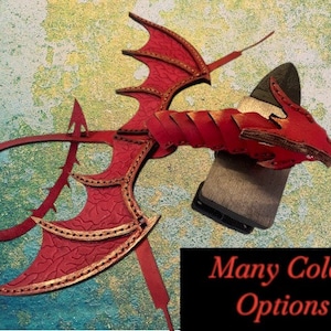 May include: Red leather dragon wings with gold accents and a tail. The wings are attached to a leather strap. The dragon wings are a decorative piece for a costume or cosplay.
