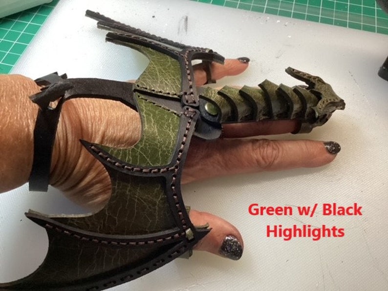 May include: Green leather dragon claw ring with black highlights. The ring is worn on a finger and has a wrist cuff.